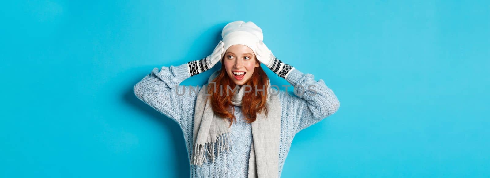 Winter and holidays concept. Happy redhead girl in beanie hat, scarf and gloves looking right and smiling, standing against blue background.