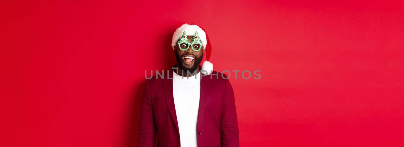 Merry Christmas. Cheerful Black man wearing funny party glasses and santa hat, smiling joyful, celebrating winter holidays, standing over red background by Benzoix