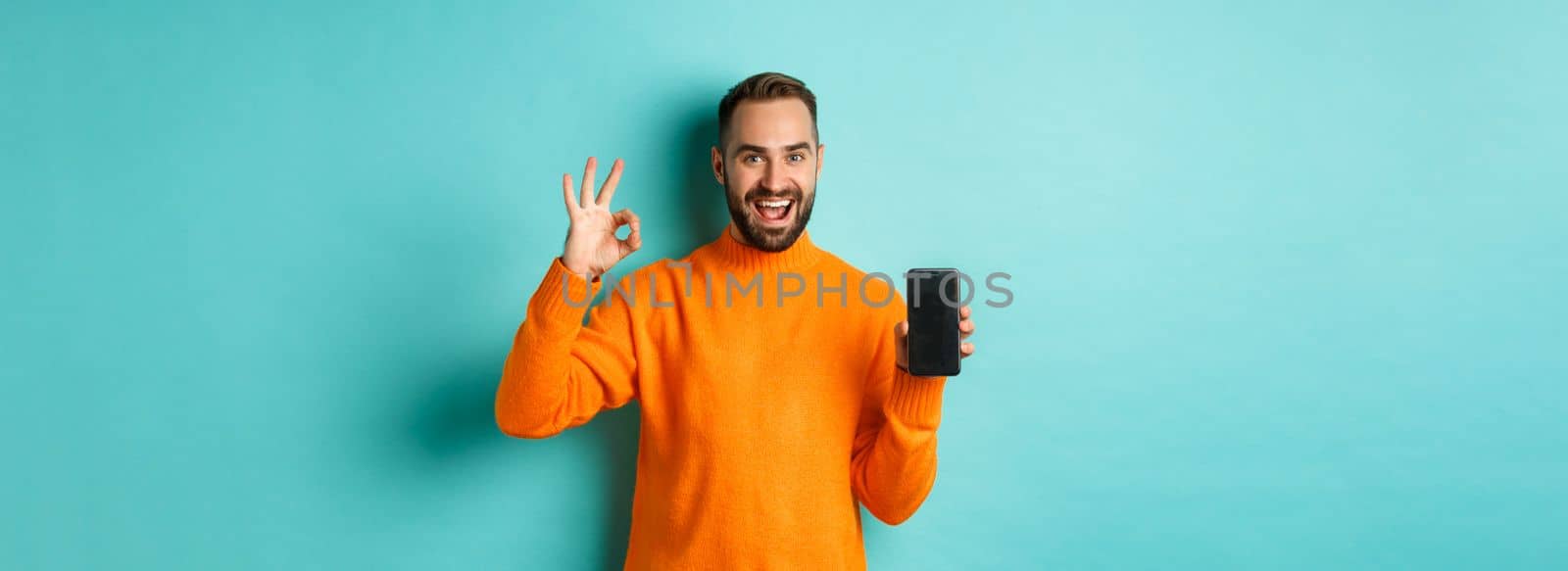 Photo of caucasian man showing mobile screen and okay sign, approve online store, smartphone app, standing satisfied over light blue background by Benzoix