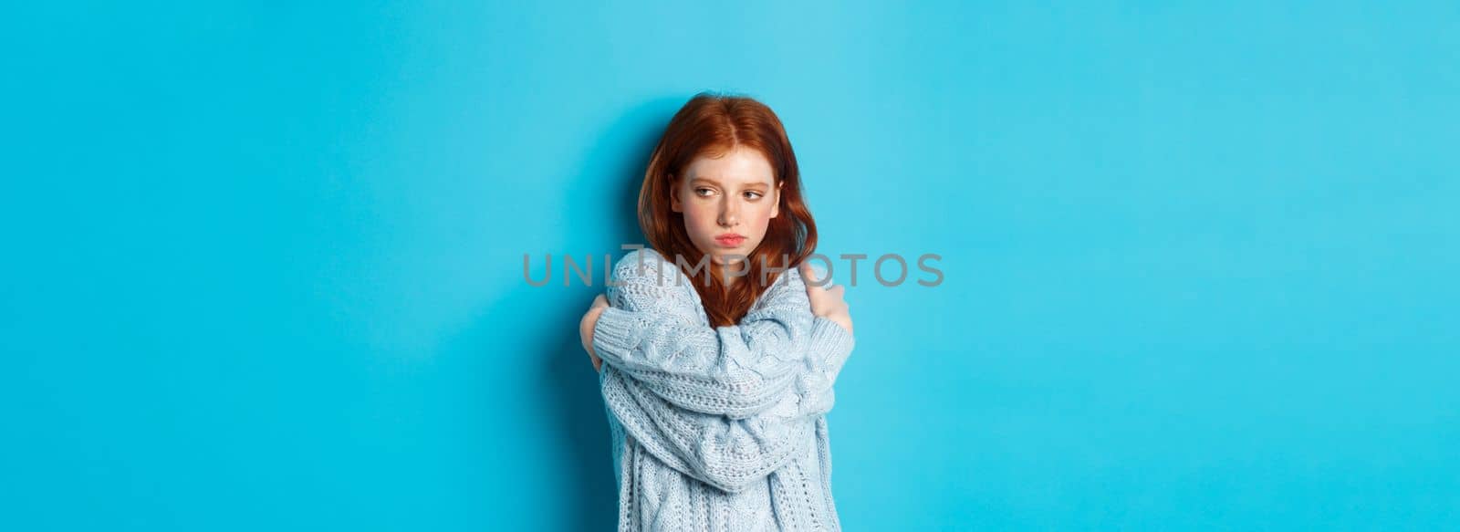 Silly and cute redhead girl pucker lips and looking offended, hugging herself and looking away offended, sulking and being upset, standing over blue background.