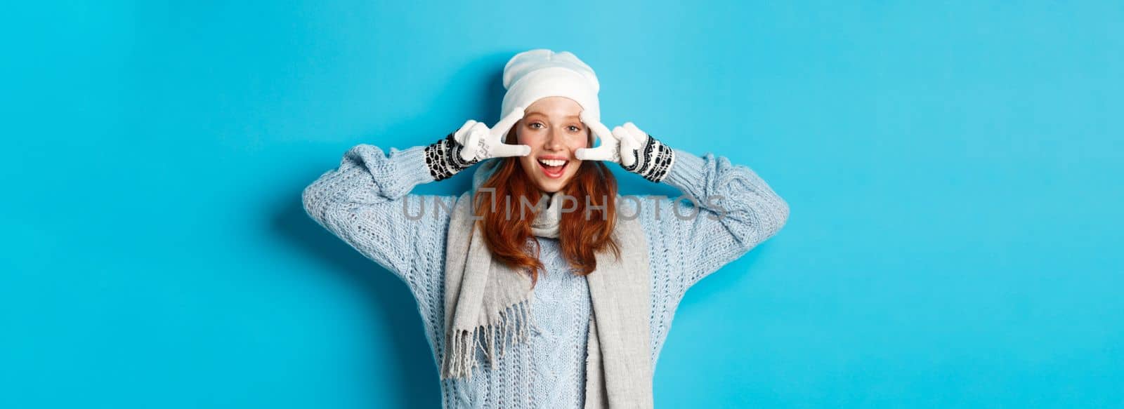 Winter and holidays concept. Cute redhead teen girl in beania, gloves and sweater showing peace sign, looking left at camera and wishing merry christmas, standing against blue background.
