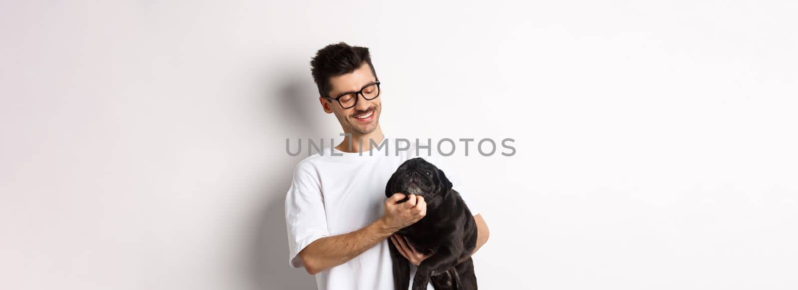 Happy dog owner scratching cute black pug face and smiling, pet his animal, standing over white background.