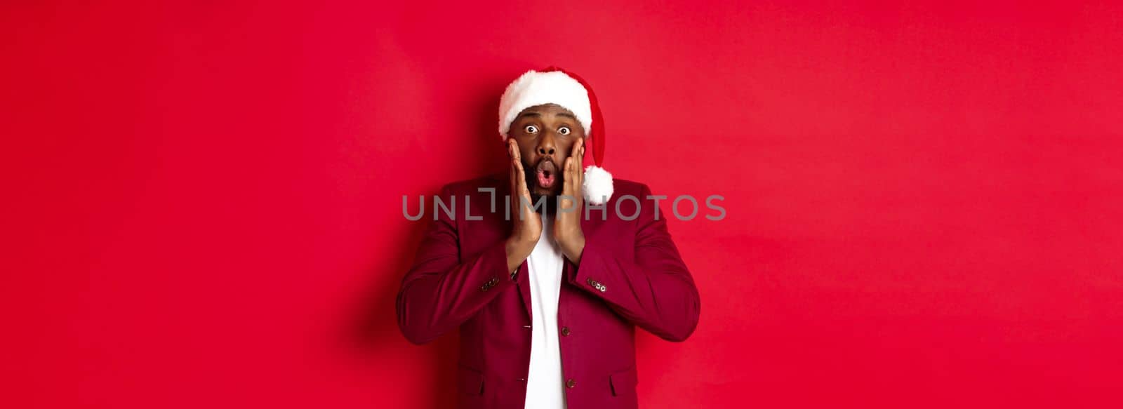 Christmas, party and holidays concept. Shocked Black man in santa hat reacting to new year offer, standing against red background.