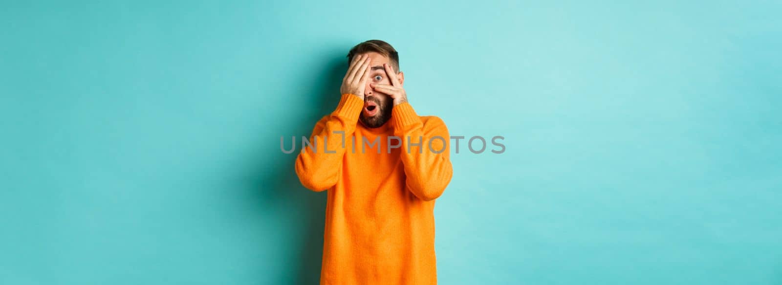 Curious man peeking at something, shut eyes but looking through fingers with amazement and interest, standing over light blue background.