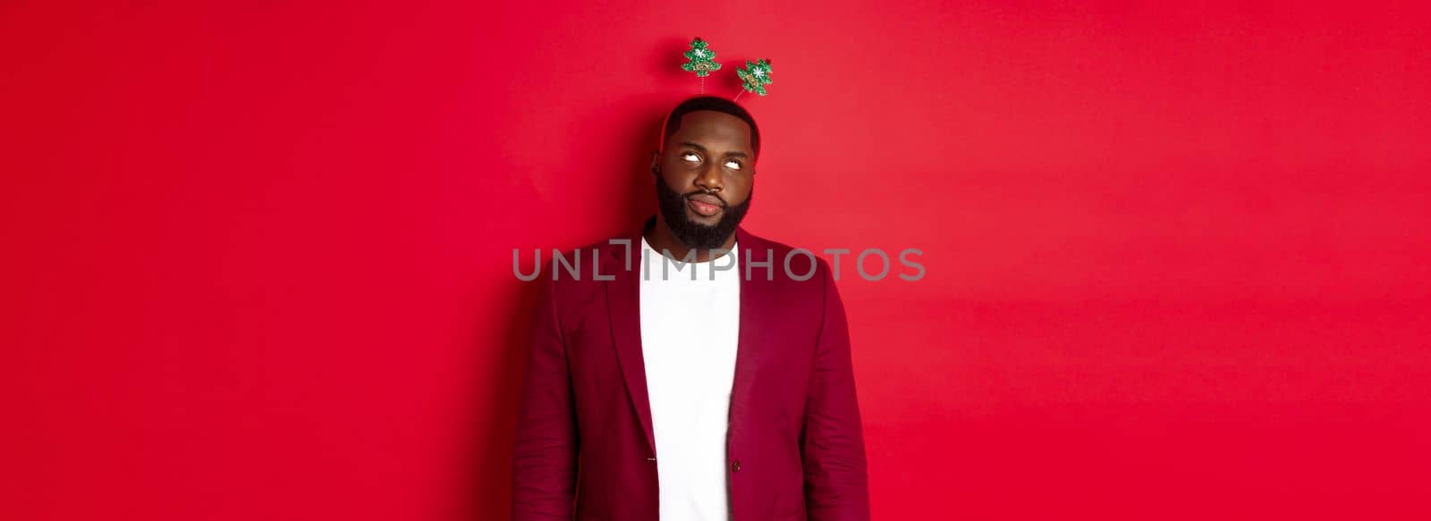Merry Christmas. Annoyed african american man roll eyes, wearing silly headband on party, standing over red background.