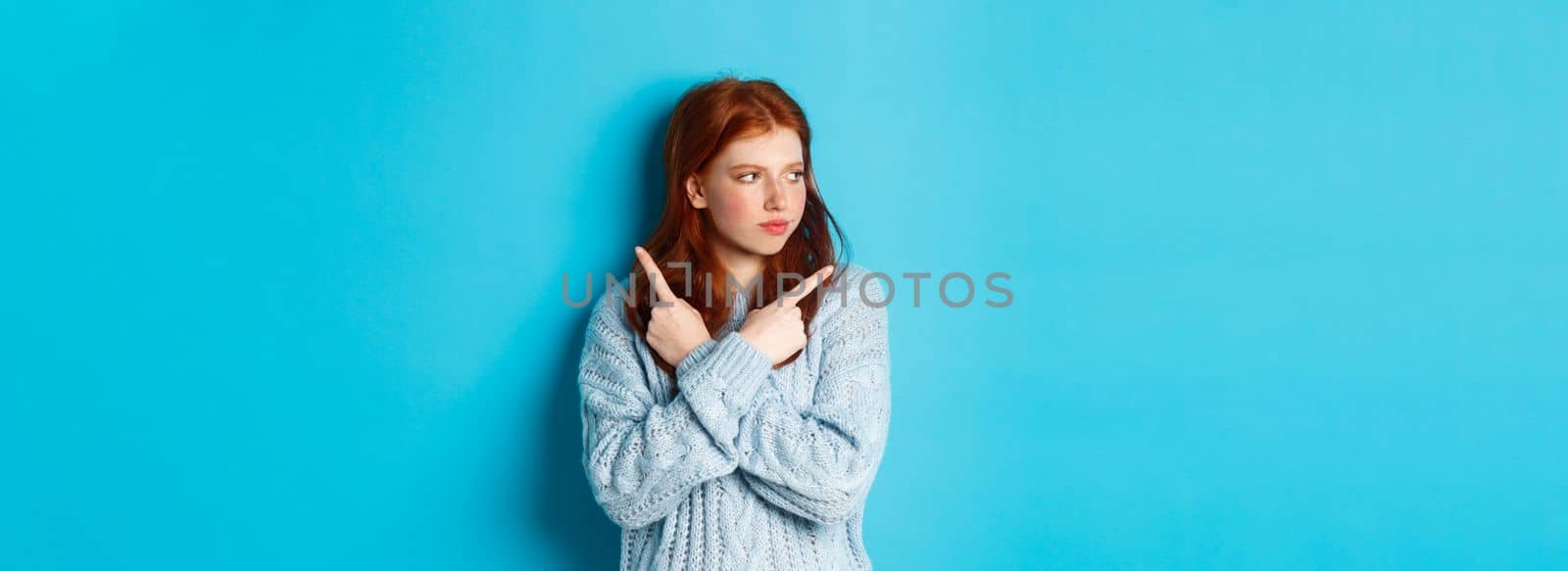 Indecisive redhead teenage girl making decision, pointing fingers sideways and looking left doubtful, standing in sweater against blue background.