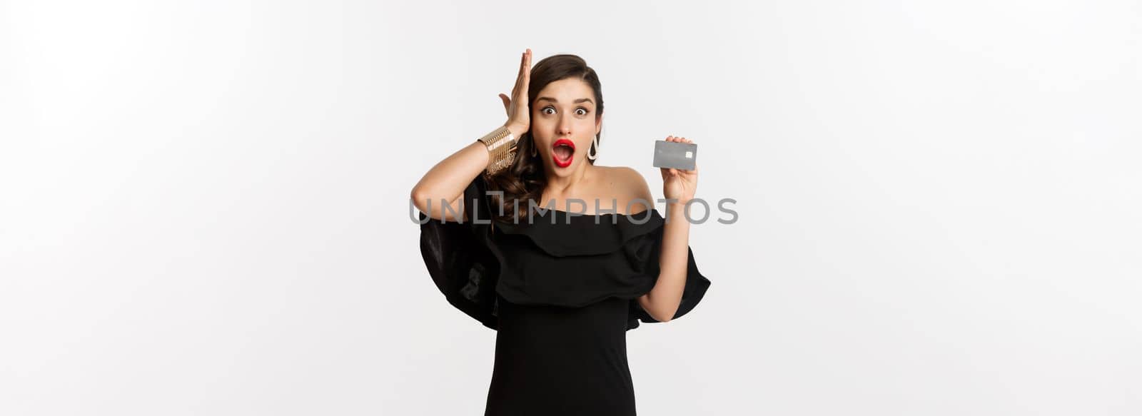 Fashion and shopping concept. Shocked woman in black dress showing credit card, looking in awe at camera, white background.