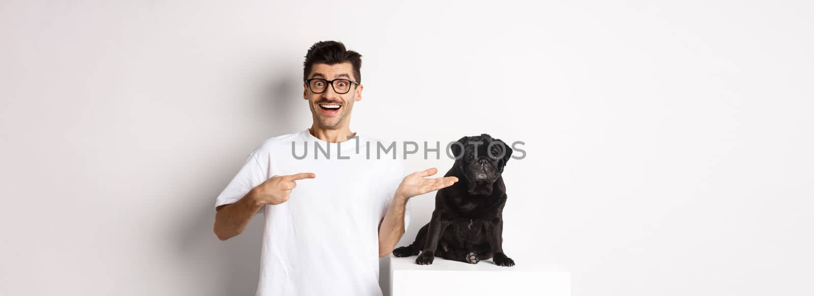 Cheerful young man pointing finger at his dog, showing small cute black pug sitting, white background.