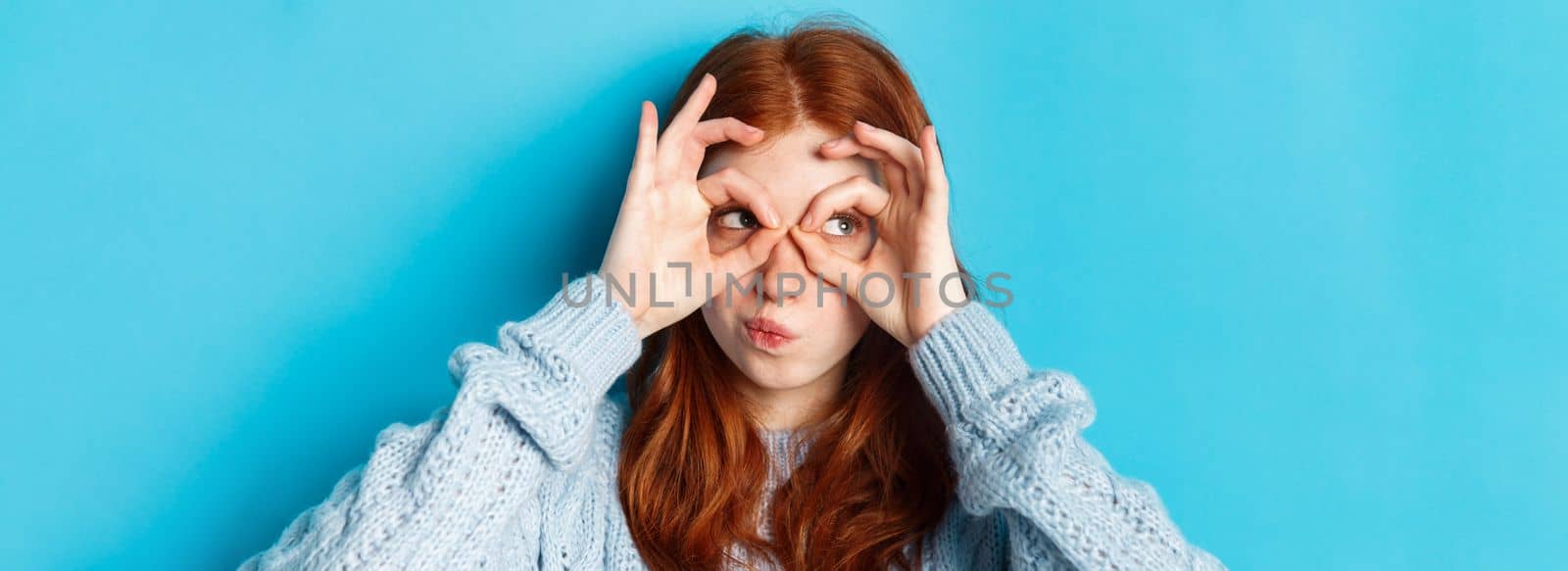 Headshot of funny redhead girl thinking, looking through finger glasses with thoughtful face, standing against blue background by Benzoix