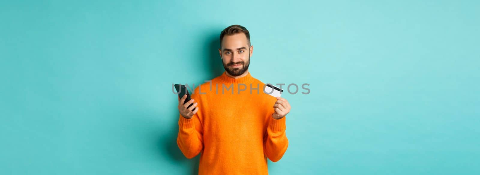 Online shopping. Handsome caucasian man in orange sweater, using credit card and mobile phone, standing over light blue background by Benzoix