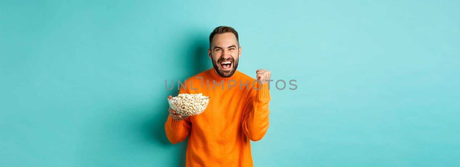 Cheerful handsome man in orange sweater, saying yes, cheering and rejoicing, eating popcorn and watching sports, making fist pump satisfied, standing over blue background.