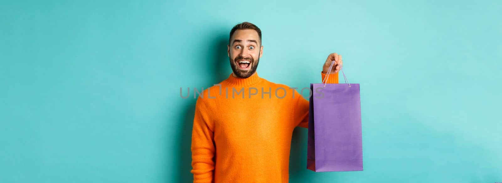 Excited adult man holding purple shopping bag and smiling, going to mall, standing over turquoise background by Benzoix