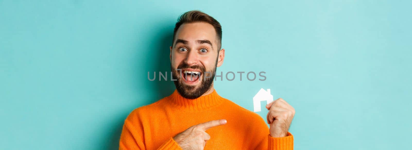 Real estate. Excited bearded man pointing at small paper house maket, standing amused over light blue background.