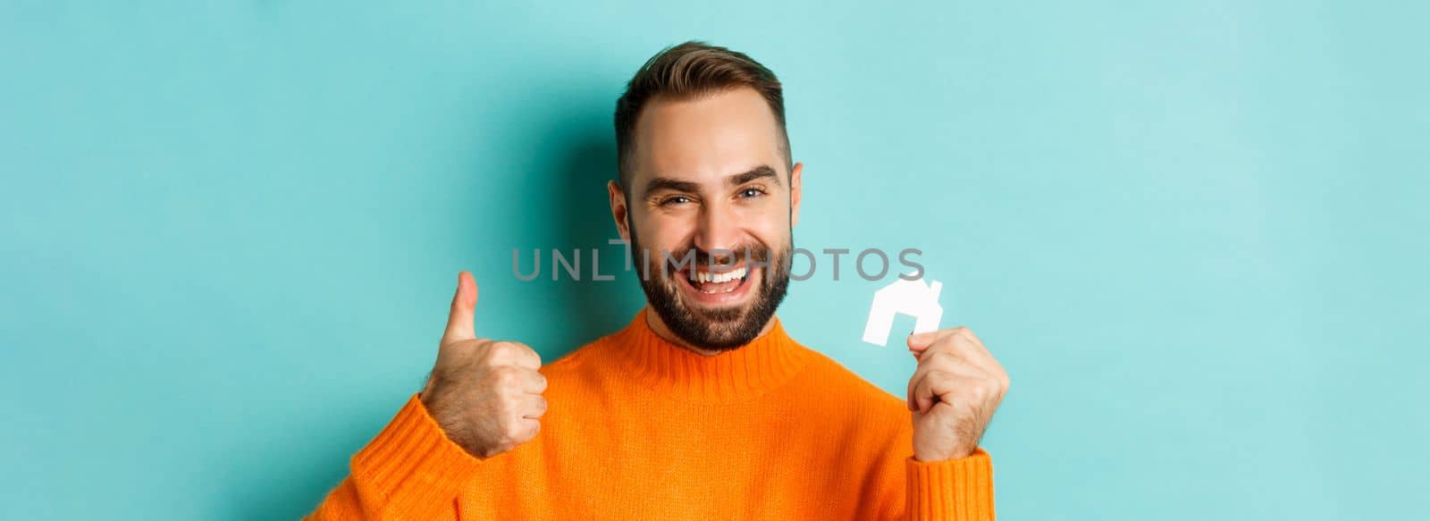Real estate. Close-up of satisfied man showing thumb-up and small paper house maket, smiling pleased, standing over light blue background by Benzoix