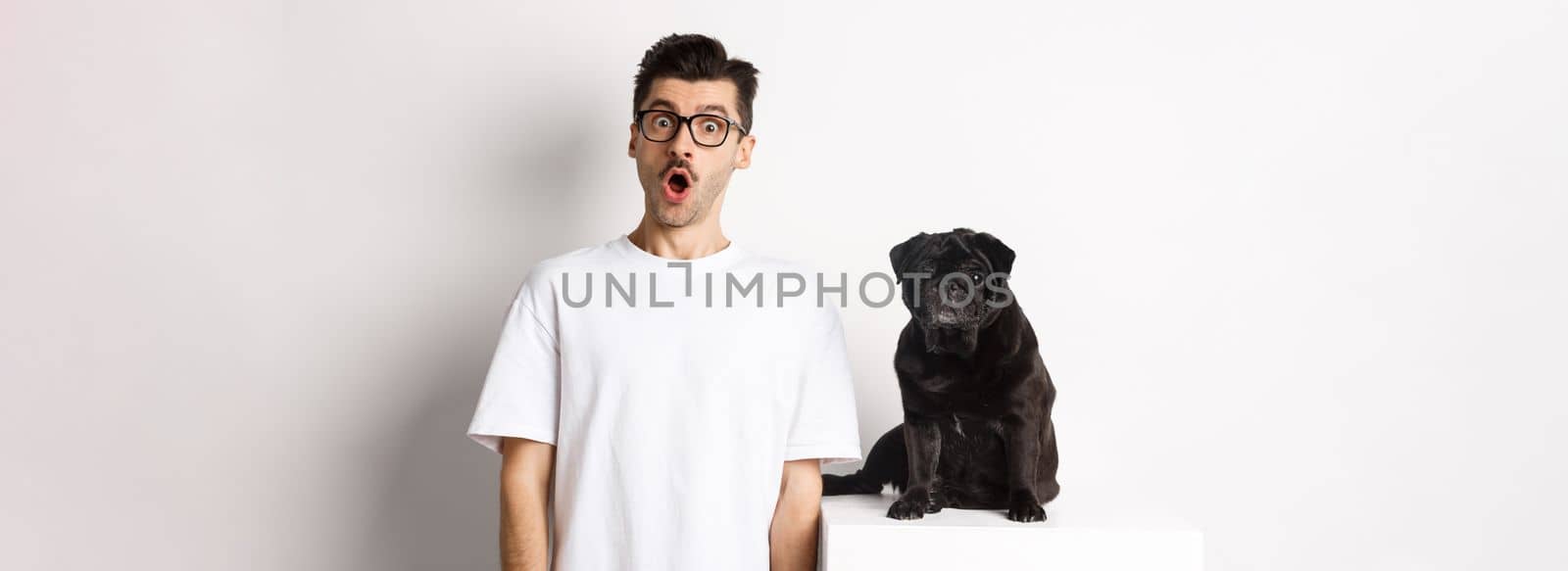 Image of dog owner and cute black pug looking at camera surprised and amazed, standing over white background.