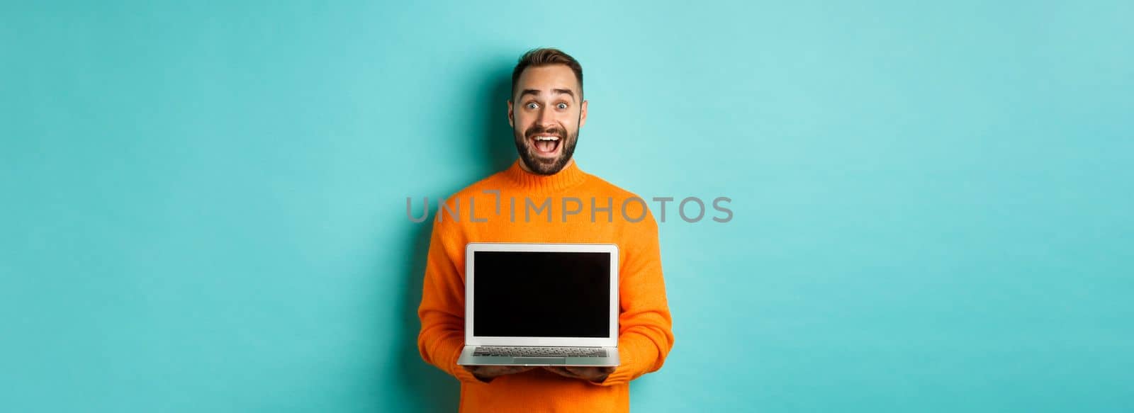 Handsome bearded man in orange sweater showing laptop screen, demonstrating promo, standing over light blue background.