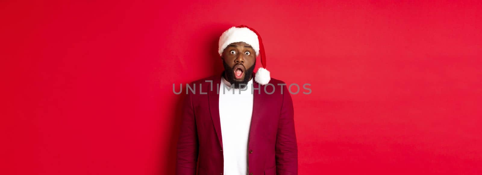 Christmas, party and holidays concept. Shocked Black man in santa hat drop jaw and stare at camera, stand against red background.