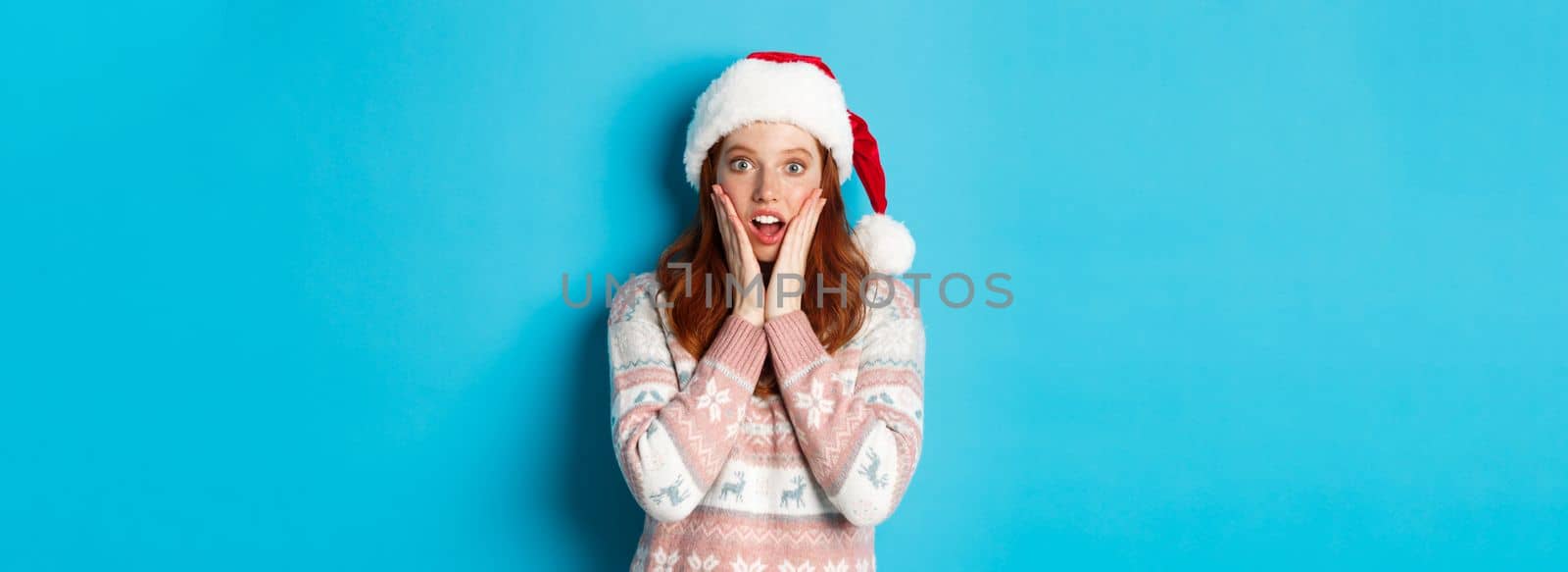Winter and Christmas Eve concept. Impressed redhead girl in santa hat hear amazing news, touching cheeks and open mouth fascinated, standing against blue background by Benzoix