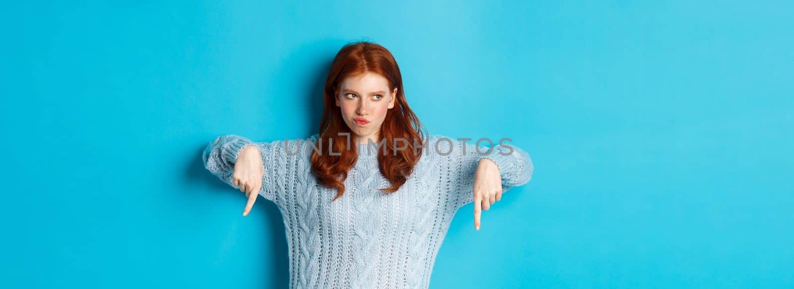 Winter holidays and people concept. Indecisive redhead girl in sweater pointing fingers down and thinking, having doubts, standing over blue background.