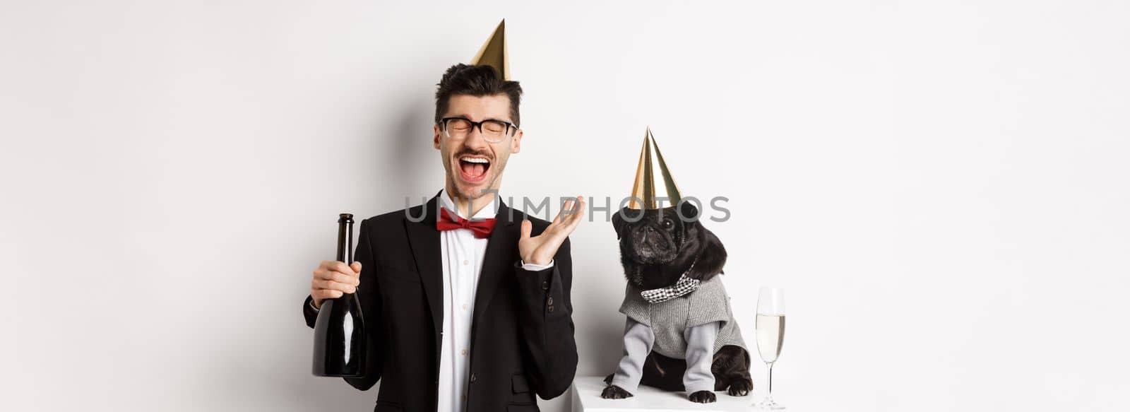 Happy young man celebrating holiday with cute dog, holding champagne and smiling, pug and owner wearing party costumes, white background.