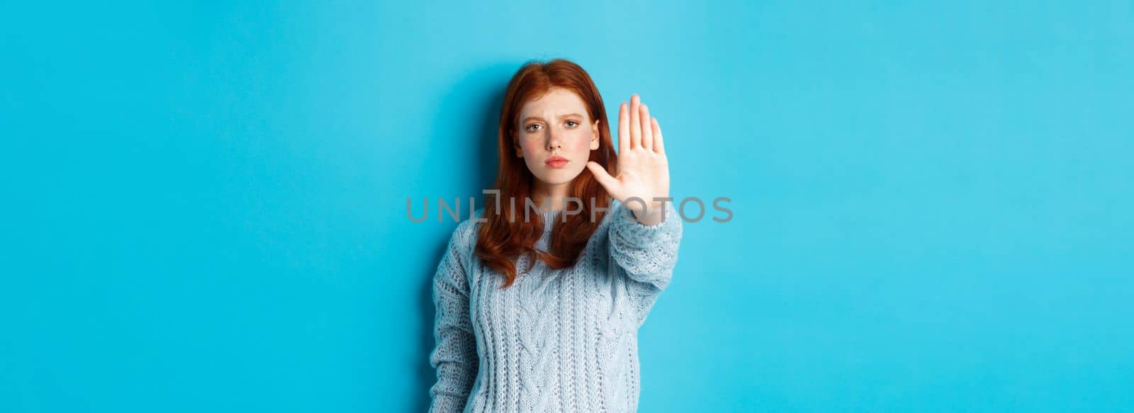 Serious and confident redhead girl telling to stop, saying no, showing extended palm to prohibit action, standing over blue background by Benzoix