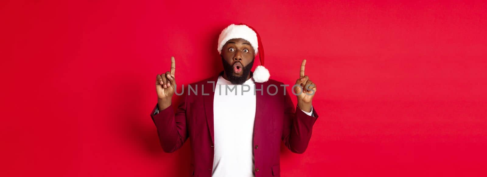 Christmas, party and holidays concept. Impressed Black man showing offer, pointing fingers up at logo, saying wow, standing against red background.