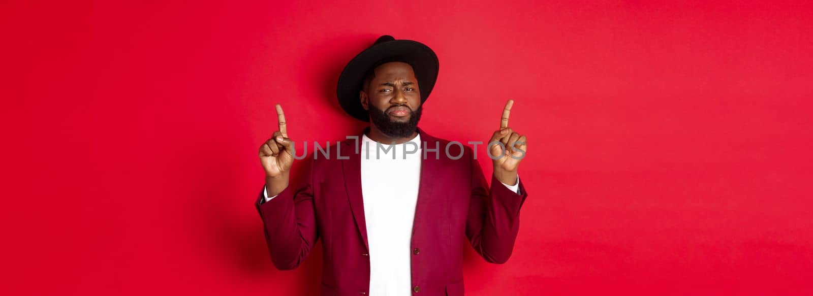 Winter holidays and shopping concept. Skeptical Black man grimasing and showing dislike, pointing fingers up at bad offer, standing over red background.