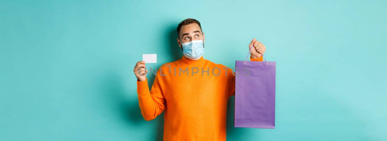 Covid-19, pandemic and lifestyle concept. Thoughtful man in face mask, holding purple shopping bag and credit card, thinking or imaging, standing over turquoise background.