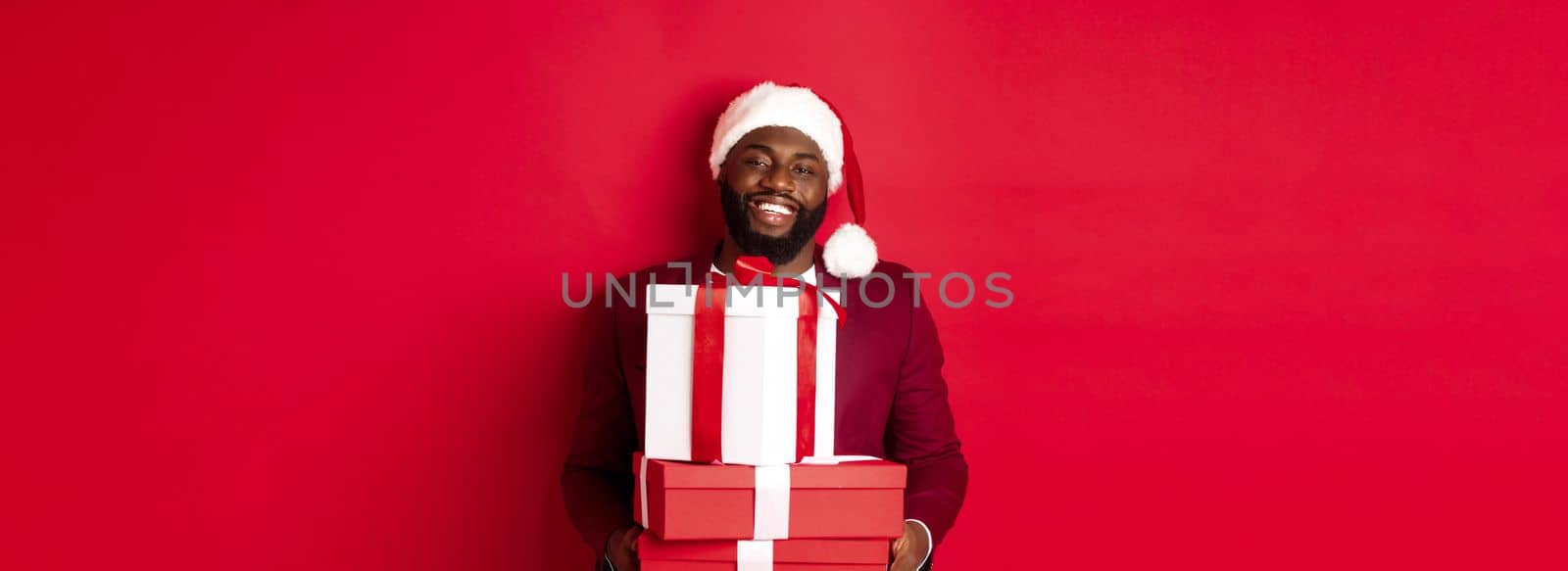 Christmas, New Year and shopping concept. Happy Black man in santa hat and blazer holding xmas presents, bring gifts and smiling, standing against red background by Benzoix