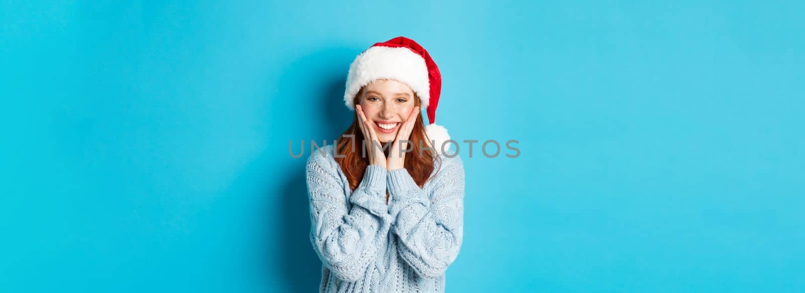 Winter holidays and Christmas Eve concept. Cheerful redhead girl in santa hat, celebrating New Year, blushing and smiling happy, standing over blue background.