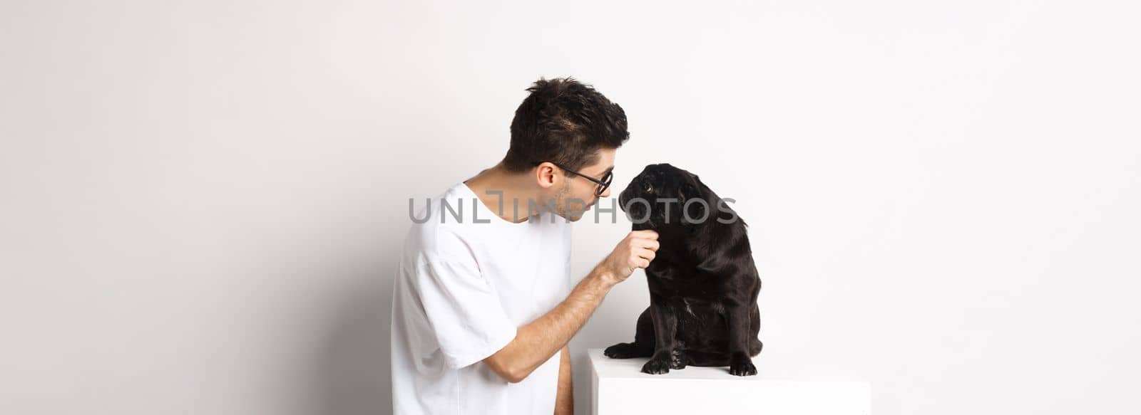 Handsome young hipster in glasses, pet his dog, looking at each other, standing over white background.