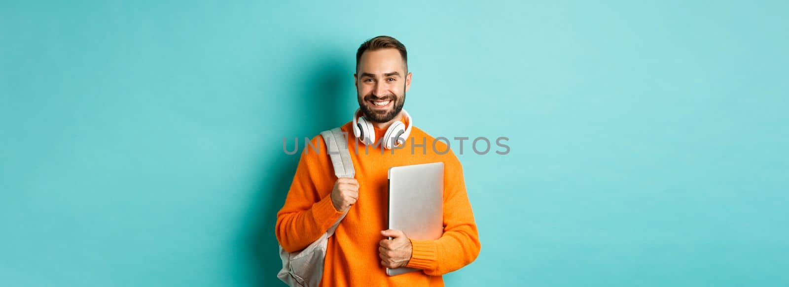 Happy man with backpack and headphones, holding laptop and smiling, going to work, standing over turquoise background.