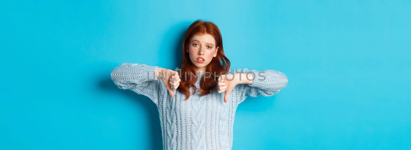 Bored and skeptical redhead girl showing thumbs down, looking unamused and uninterested, standing over blue background.