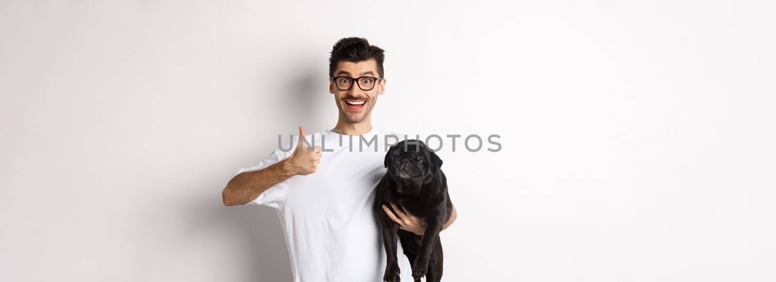 Cheerful handsome man holding dog and showing okay sign, approve or recommend product. Hipster guy carry cute black pug and looking satisfied, white background by Benzoix