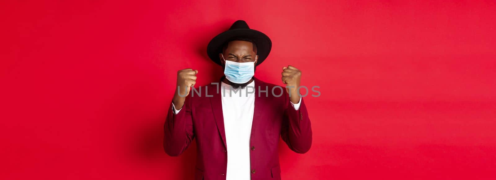 Covid-19, quarantine and holidays concept. Cheerful african american man showing clenched fist and rejoicing of winning, achieve goal, wearing medical mask from coronavirus.