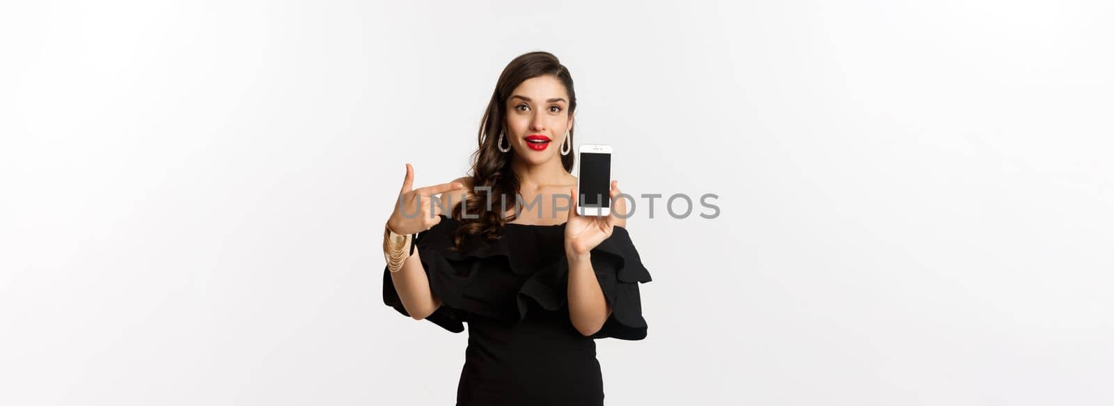 Online shopping concept. Fashionable woman in black dress pointing finger at smartphone screen, showing application, standing over white background by Benzoix