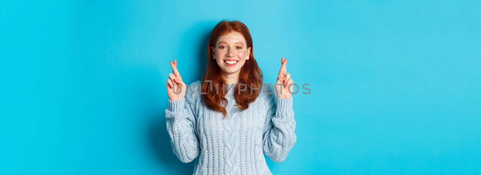 Hopeful redhead girl making a wish, cross fingers for good luck, smiling and anticipating good news or positive result, standing against blue background.