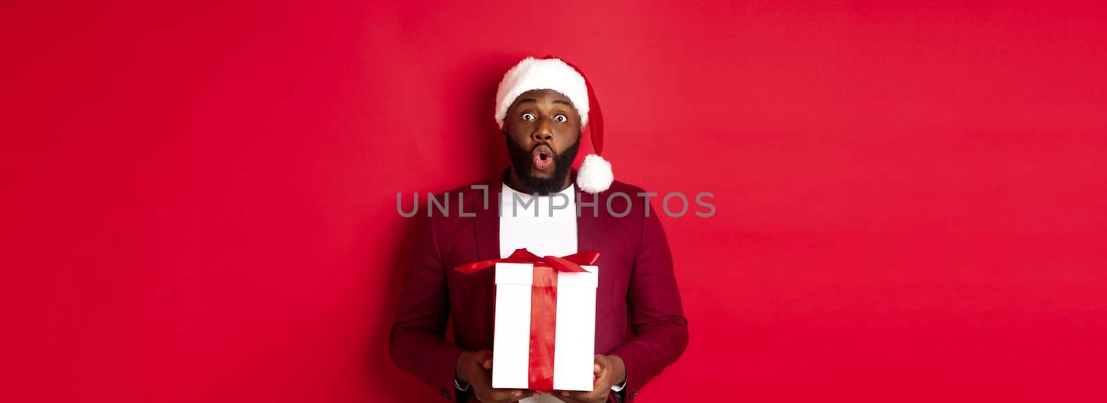 Christmas, New Year and shopping concept. Surprised african american man in santa hat saying wow, holding holiday gift and looking at camera amazed, red background by Benzoix