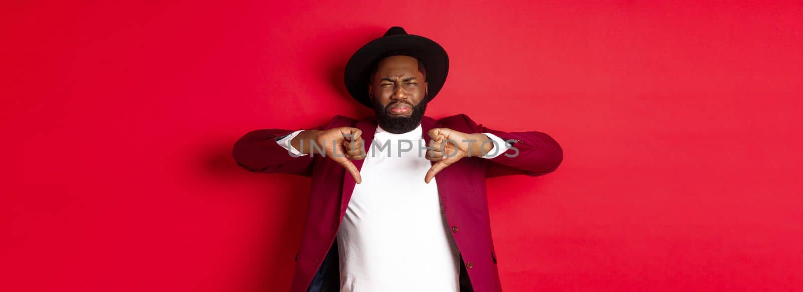 Disappointed Black man leave negative feedback, showing thumb down and grimacing from dislike and disgust, standing in party clothes over red background by Benzoix