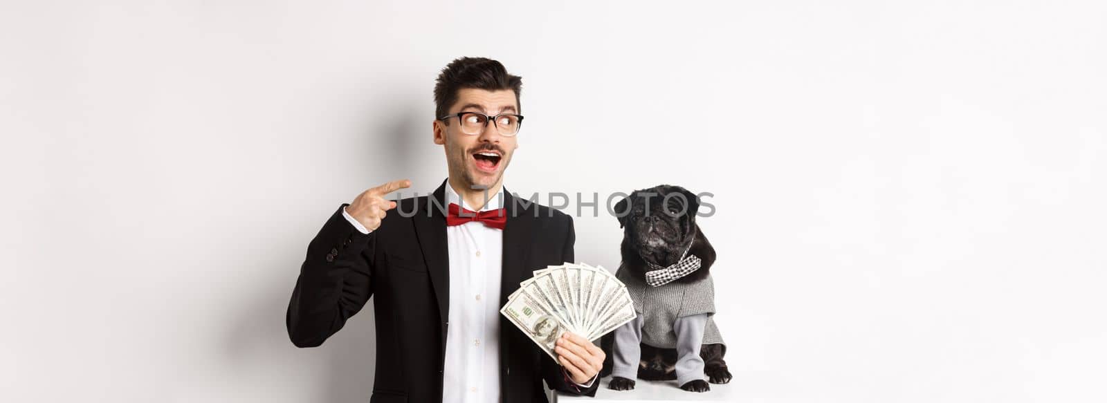 Happy young man in suit earn money with his dog. Guy rejoicing, holding dollars and pointing left, black pug in costume staring at camera, white background by Benzoix