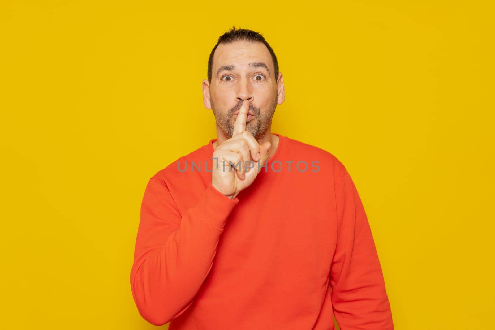 A serious handsome man in a red sweater, puts his index finger to his lips, tries to keep the conspiracy, looks to the side and says: Shh, be quiet please. Isolated shot of man shows silence sign
