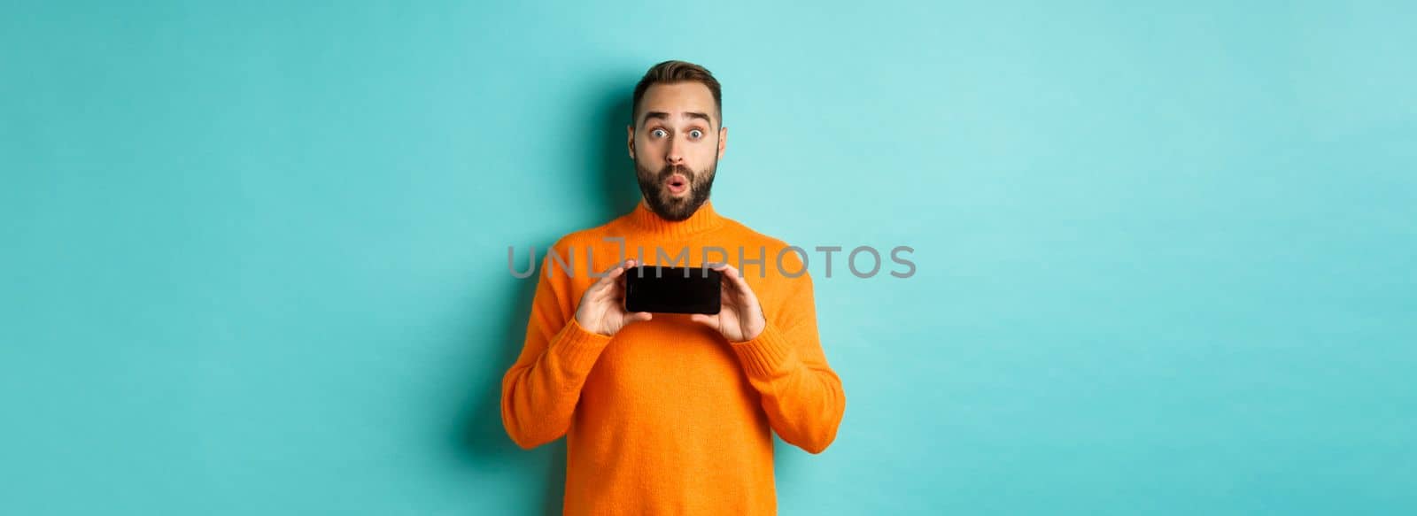 Impressed man showing smartphone screen, stare at camera amazed, demonstrate display, standing over light blue background.