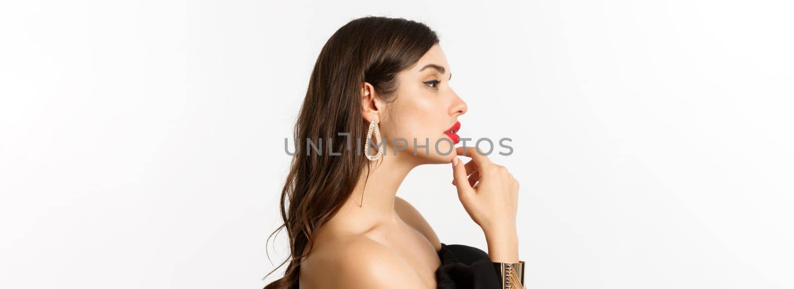 Fashion and beauty concept. Profile view of attractive and stylish woman in black evening dress, makeup and earrings, looking left sensual, standing over white background.