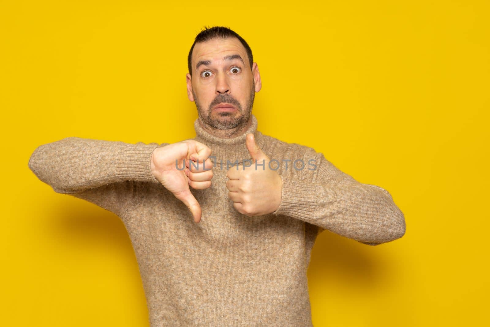Bearded hispanic man wearing a turtleneck sweater giving a thumbs up and thumbs down in concept of doubt and indecision, isolated over yellow background