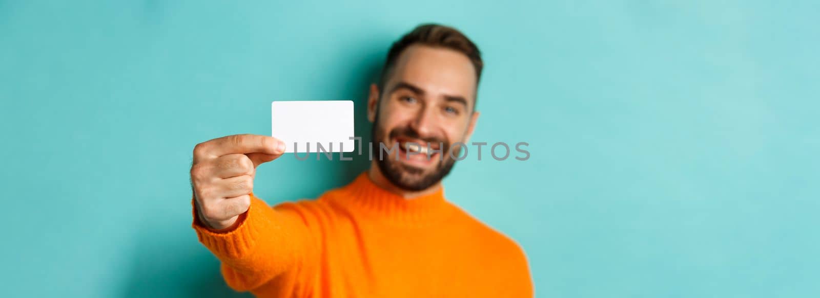 Close-up of handsome caucasian man going on shopping, showing credit card and smiling, standing over turquoise background by Benzoix
