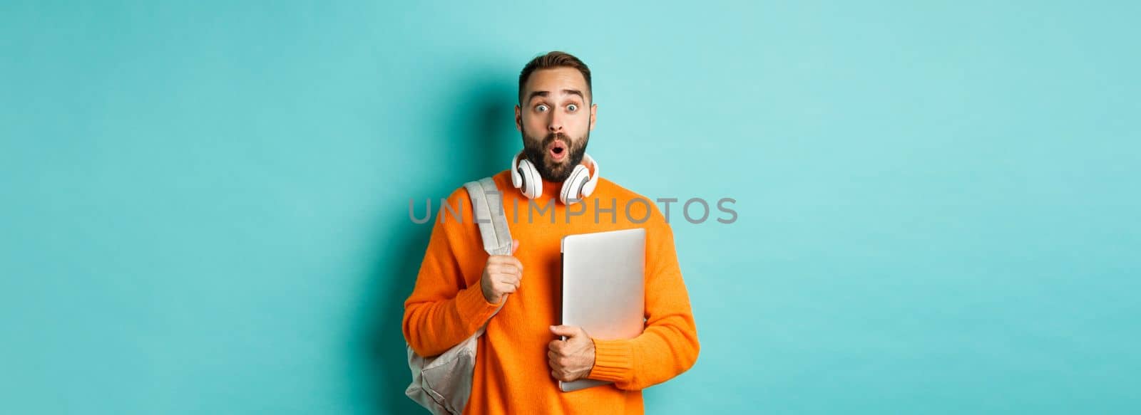 Happy man with backpack and headphones, holding laptop and smiling, looking surprised, standing over turquoise background.