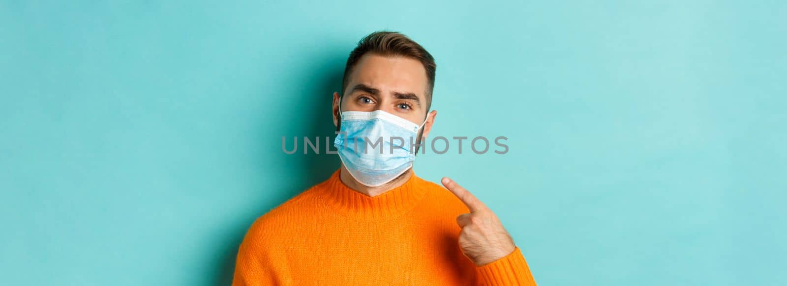 Covid-19, social distancing and quarantine concept. Upset man in complaining on face mask, pointing at it and looking bothered, standing over light blue background.