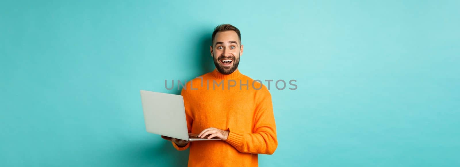 Excited smiling man working on laptop, staring at camera happy, standing in orange sweater against turquoise background by Benzoix