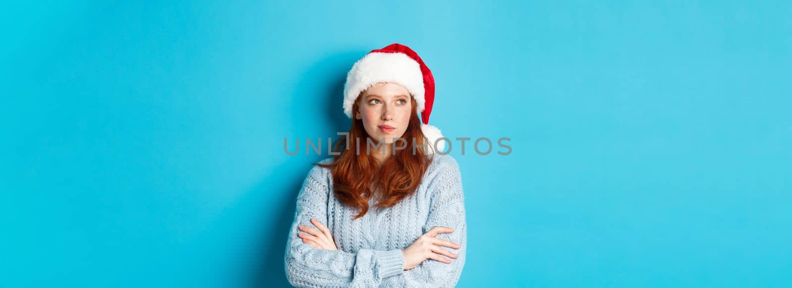 Winter holidays and Christmas Eve concept. Thoughtful redhead woman in Santa hat and sweater, looking left and pondering, making xmas plans, standing over blue background.