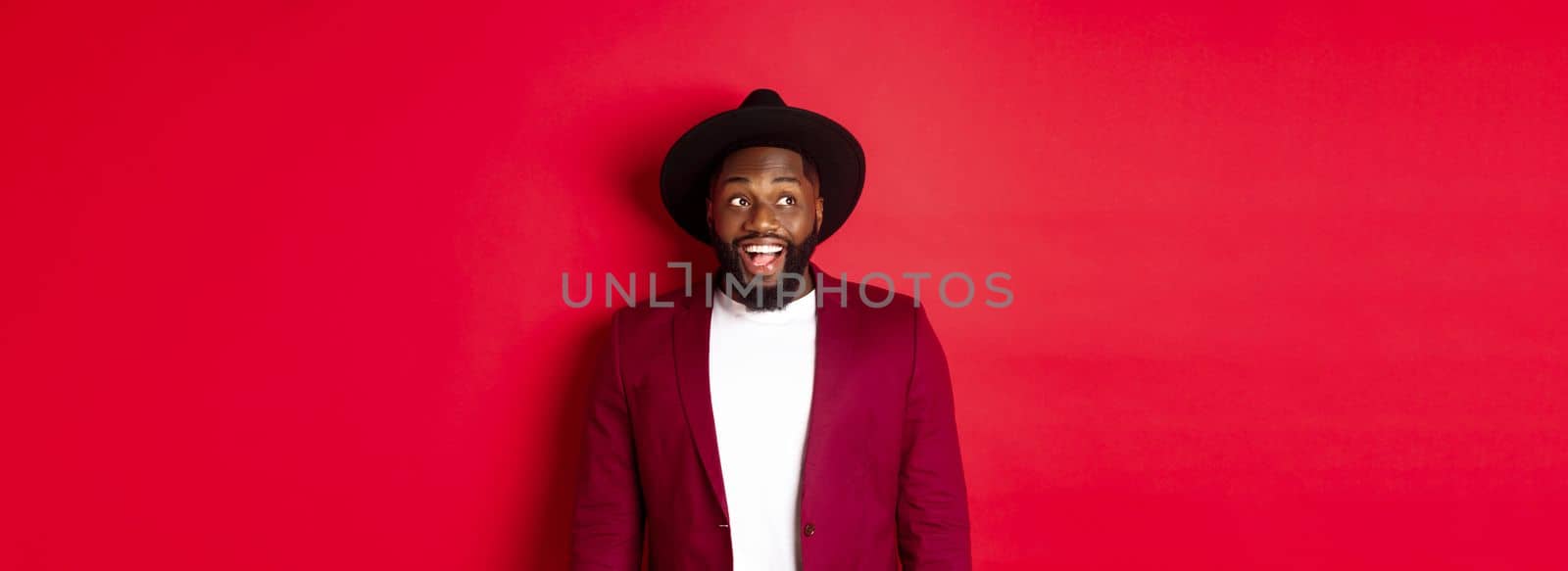 Fashion and party concept. Excited african american man looking at logo, staring upper left corner with happy smile, standing against red background by Benzoix